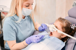 Female dentist checking child teeth with dental explorer and mirror while girl lying in dental chair with inhalation sedation at dental office. Concept of pediatric, sedation dentistry and dental care