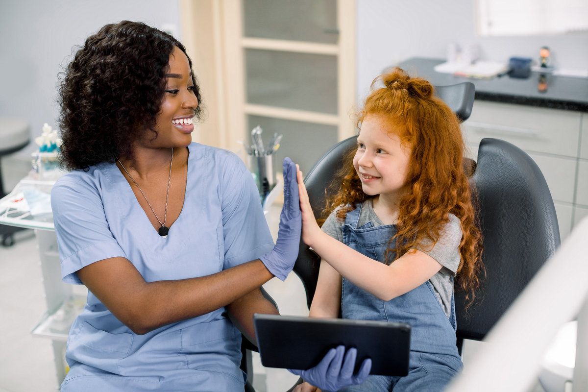 Medicine, pediatric dentistry and oral care concept. Happy female African dentist and curly red haired kid girl, having fun and giving high five, while holding tablet pc
